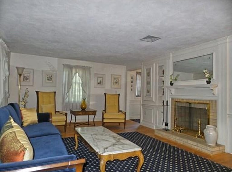 A living room with white walls, white curtains, a fireplace, large blue area rug and antique-style furniture