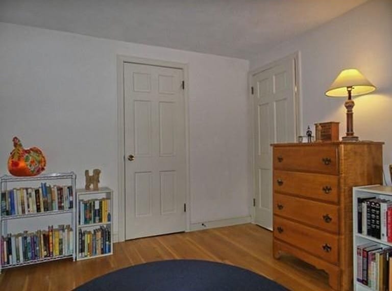 A bedroom with a wood floors and a blue rug; tall, wooden chest of drawers, and smaller white shelves with books