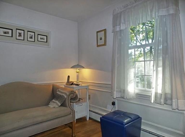 The corner of a living room near a window with white curtains, a dark blue leather ottoman, white end table and a light brown antique sofa