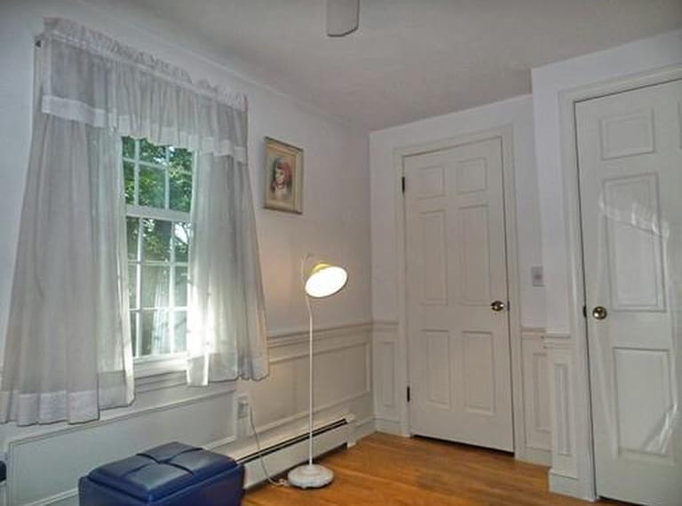 A white floor lamp near a blue leather ottoman near a window with white curtains and a door in the background