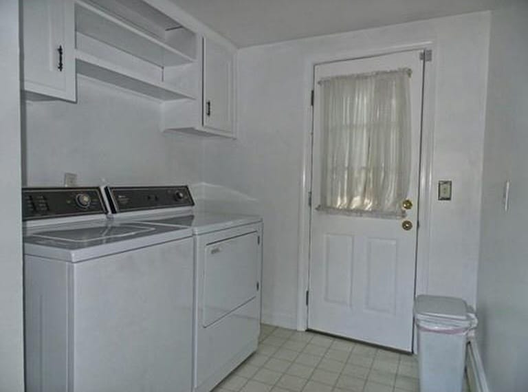 A narrow room with a white washer and dryer, shelves on top and a door with glass panes in the top half, and a white curtain