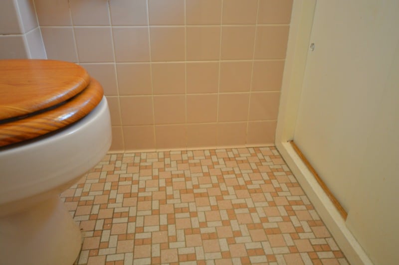 A floor with orange, pink and brown tiles, a wall with large, square pink tiles and the edge of a toilet with a wooden seat