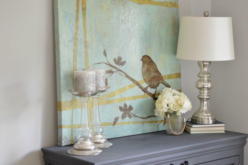 A blue/grey side table with 2 glass candle holders, a silver lamp, small vase of flowers and a large, blue and gold painting of a bird on a branch