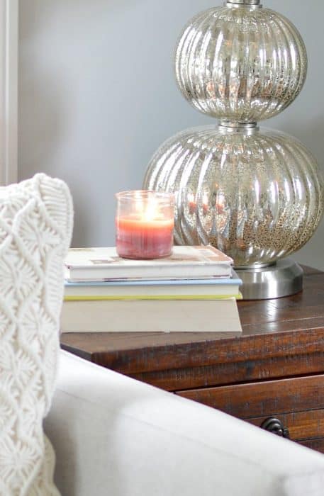 An end table with a stack of books, a peach-colored candle and a silver lamp with stacked globe base