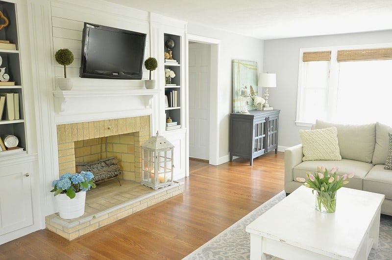 A living room with a fireplace, wall-mounted television, white coffee table and a blue cabinet in the corner by the window