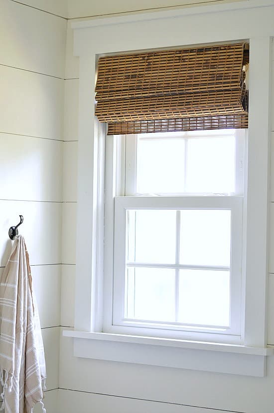 A window in the corner of a bathroom with shiplap walls, decorated with a rolled-up wooden blind