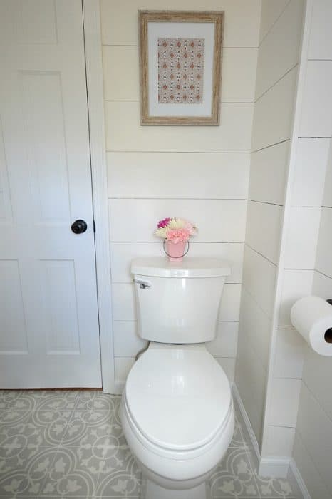 A white toilet with a short vase of flowers on top, and a tan artwork in a wood frame above