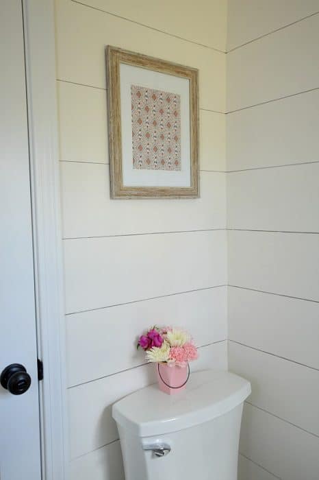 A pink vase of flowers on top of a toilet, with framed art above, on shiplap walls