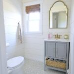 A bathroom with grey vanity, gold mirror, shiplap walls and a small window with rolled-up wooden screen