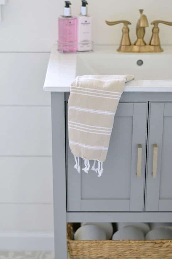 A grey bathroom vanity with a white sink and gold faucet, pink soap, tan towel and a basket below with extra toilet paper rolls