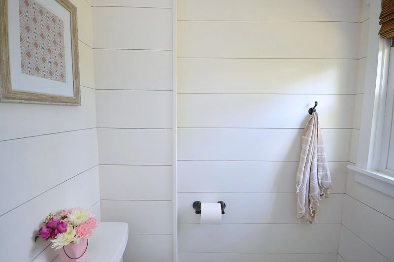 Shiplap walls with a towel hung on a dark towel hook, a roll of paper on matching hardware, and a toilet tank with flowers on top