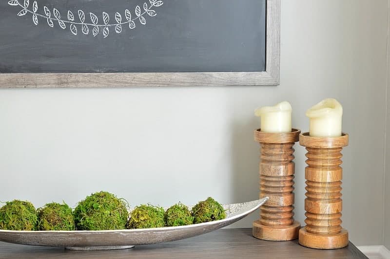 A side table with two stocky, wooden candlesticks, a narrow platter of green moss balls and the corner of a large chalkboard with chalk design