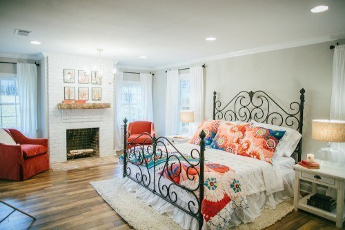 A bedroom with a fireplace and artwork above mantel, an orange chair in between two windows in the corner and a large, black iron bed with quilted bedding