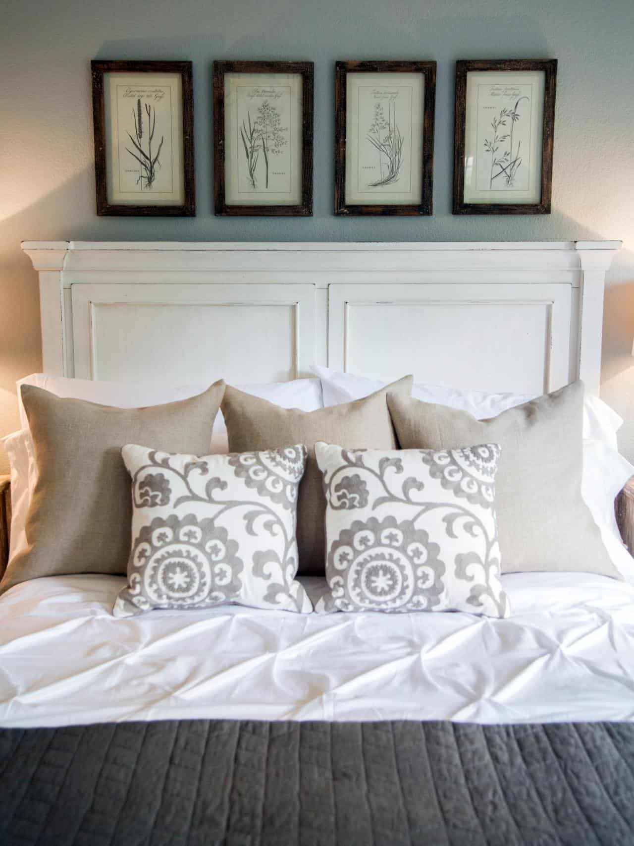 A bed with white, grey and beige bedding and pillows, a white headboard and four tall pieces of artwork above featuring botanical drawings