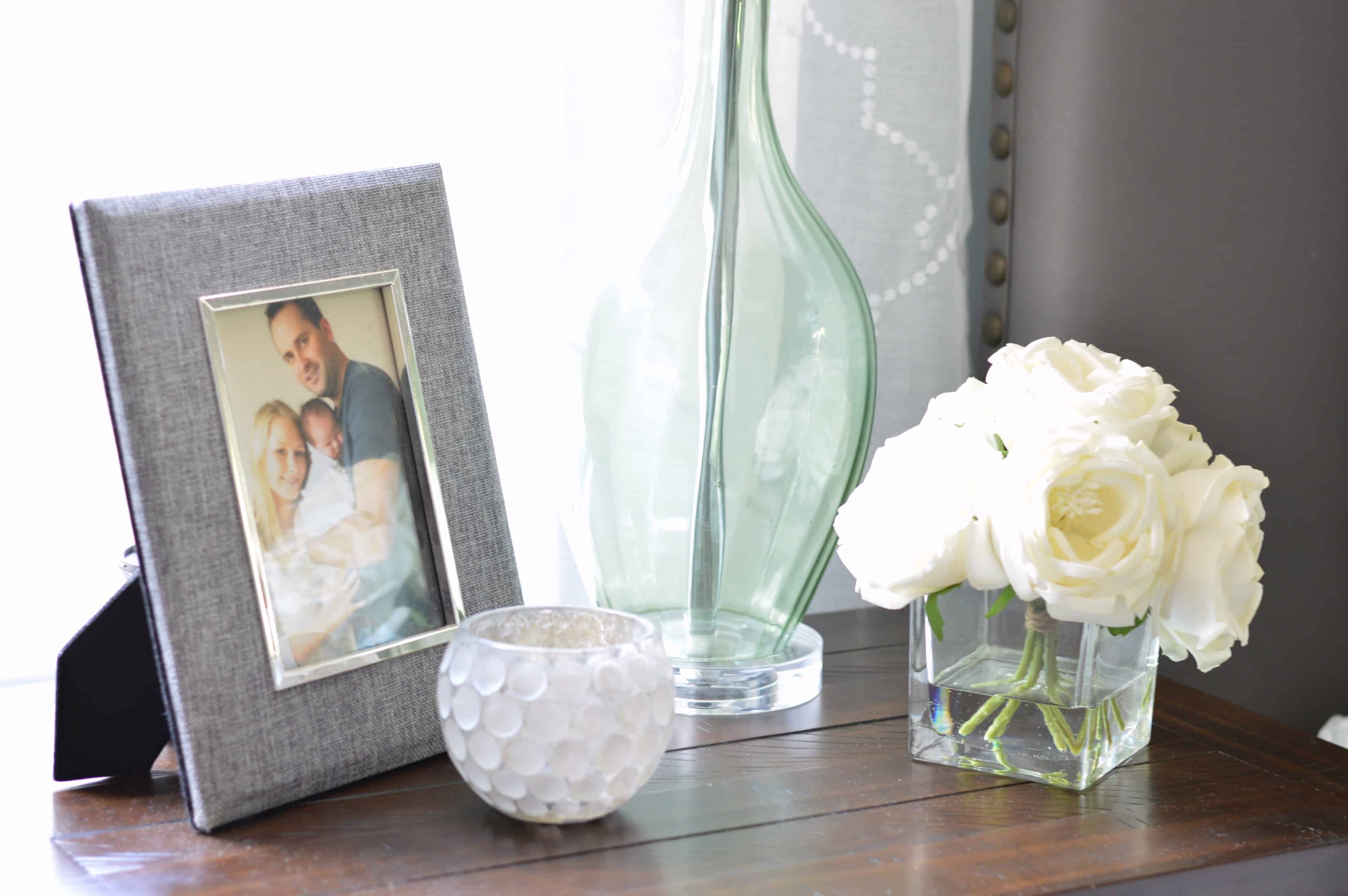 A nightstand near a window with a portrait of a family in a grey frame, a candle, the glass base of a lamp shade and a square vase with short, white flowers