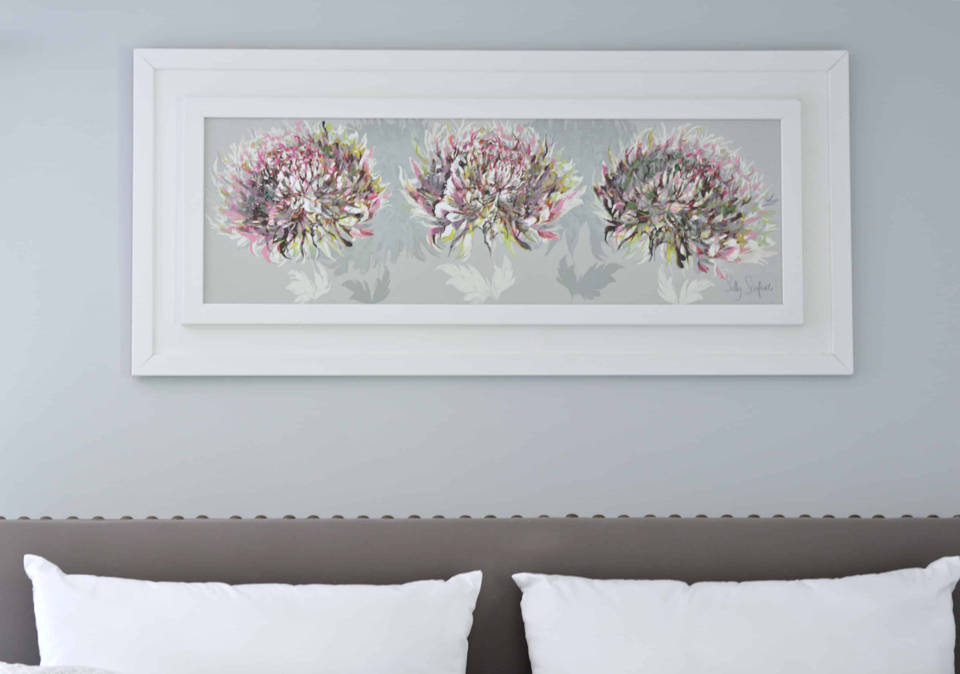 A wide, rectangular painting of three bouquets of flowers above a bed with grey, upholstered headboard 
