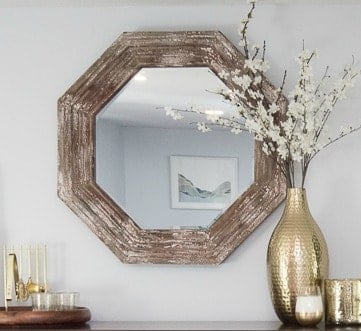 A large, hexagonal mirror with a rustic, wood frame and a large, textured gold vase with white flowers on a table below