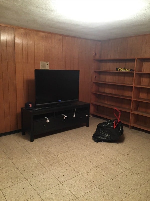 The corner of the basement with wood paneling, wooden shelves on one wall, a flat-screen television on a table and a filled garbage bag lying around