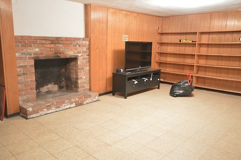 The corner of the basement with wood paneling and shelves, a table with a flat-screen television, and a brick fireplace