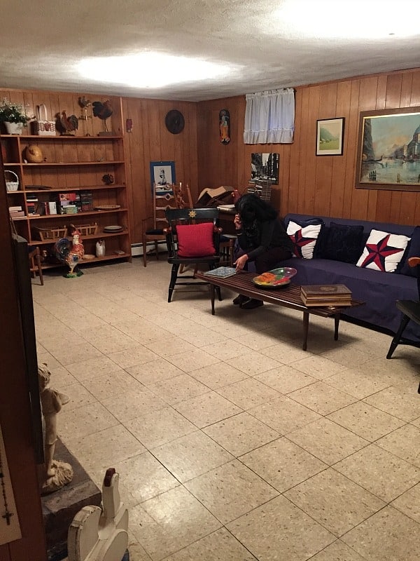 A person sitting on a navy blue couch in a basement with wood paneled walls, tile floors and miscellaneous items on a built-in shelf nearby