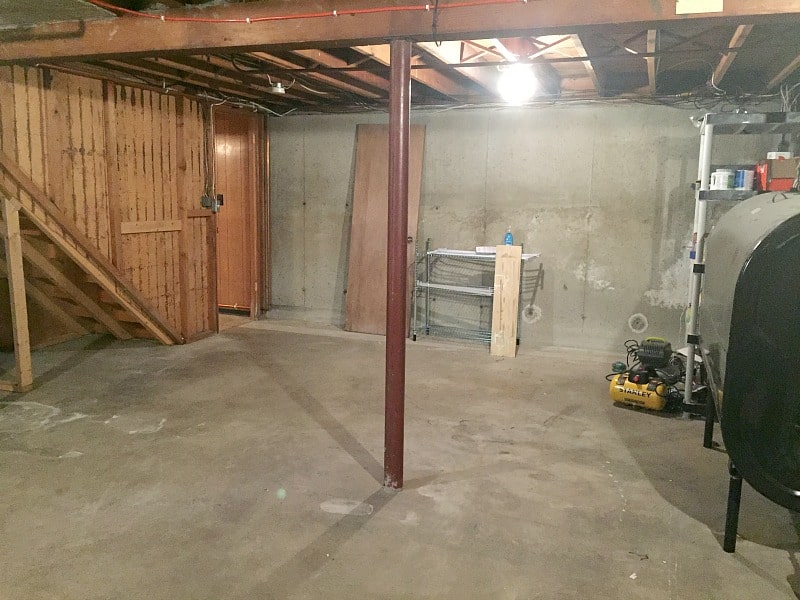 A section of the basement with concrete floors, ceiling beams exposed, a pole in the center of the room and a staircase to the left