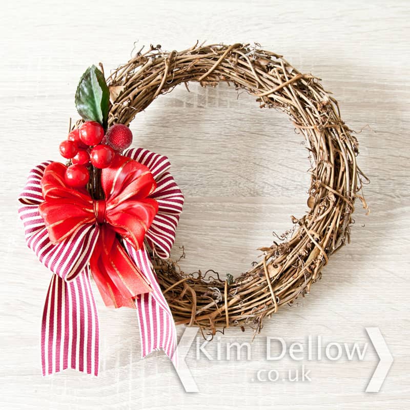 A circle of bound twigs in the shape of a wreath, with a striped red and white ribbon on the left side, topped with a smaller red ribbon and berries
