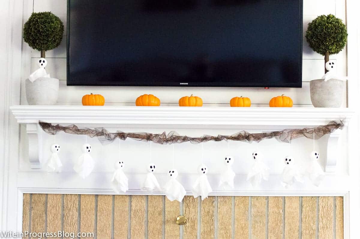 A fireplace mantel with a row of little, orange pumpkins on top and tissue ghosts hanging down from a string of tulle