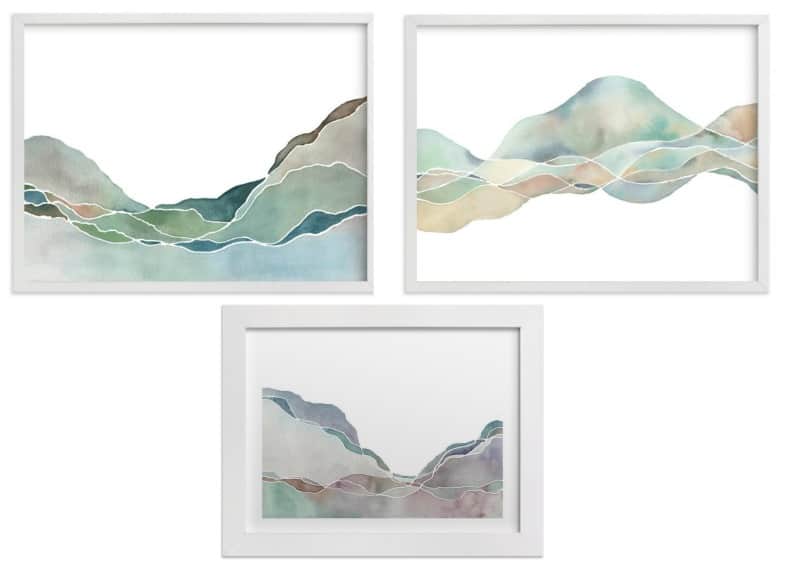 Three landscape prints of mountains in shades of blue and green, in white frames