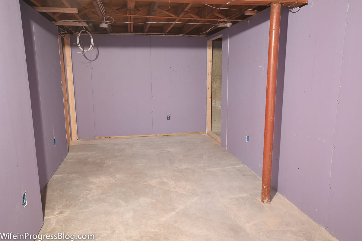A basement room with new beige carpeting, new drywall and the drop ceiling removed so the upper floor beams are exposed