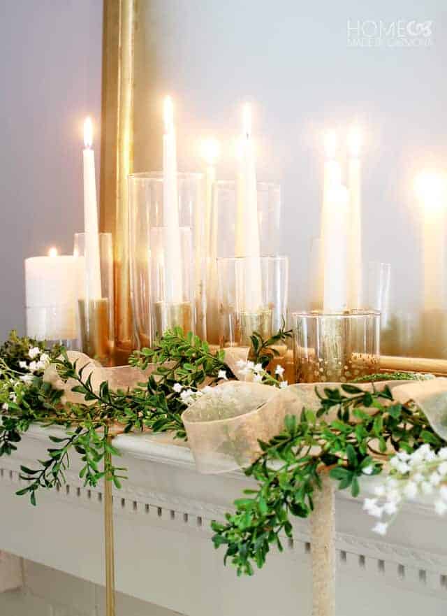 A large collection of lit candles in glass candle holders, among greenery wrapped in sheer, beige ribbon on a fireplace mantel