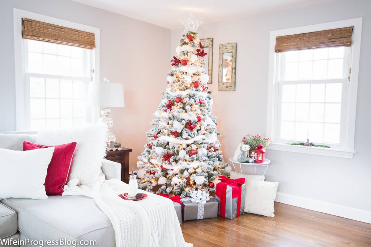 A tall Christmas tree in the corner of a living room with gifts below and a plate of milk and cookies resting on a sofa nearby