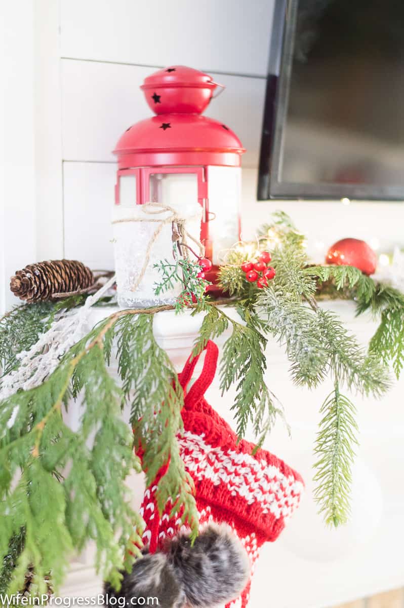 A large pine branch resting on a fireplace mantel, with a red lantern, top of a red and white stocking and pine cones