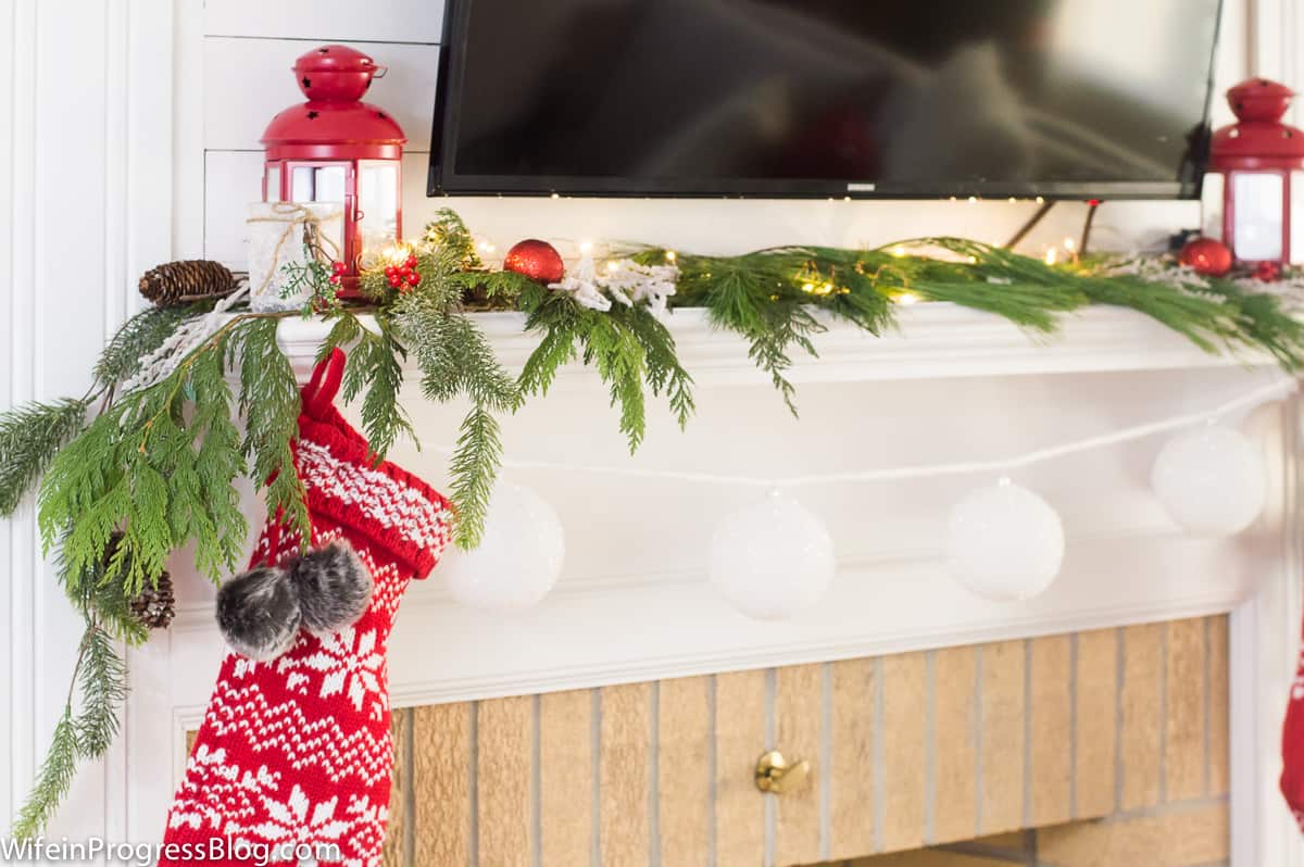 The corner of a fireplace mantel with pine branches, white and red ornaments, a red lantern and red and white stocking