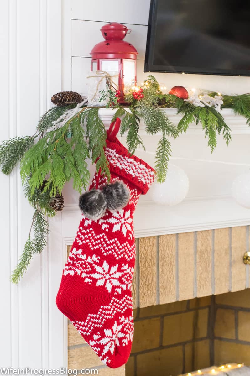 A red and white snowflake stocking hung from a white, wooden fireplace mantel and a pine branch nearby