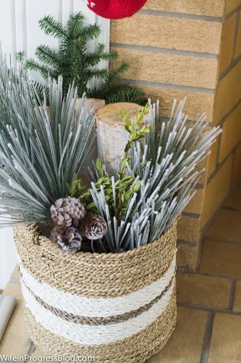 A close up of a wicker basket near a fireplace, holding firewood and decorative pine cones and greenery