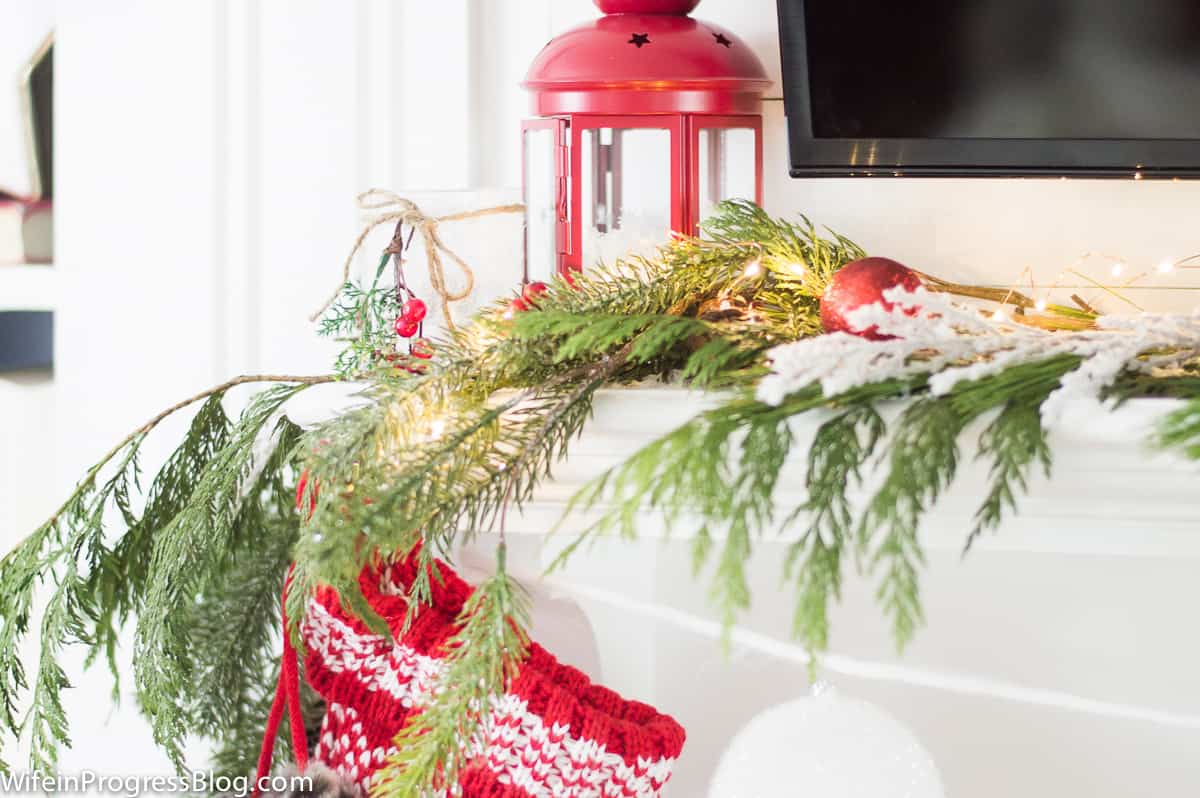 A red lantern, pine branches, white candle and a stocking hung from a fireplace mantel, below a wall-mounted television