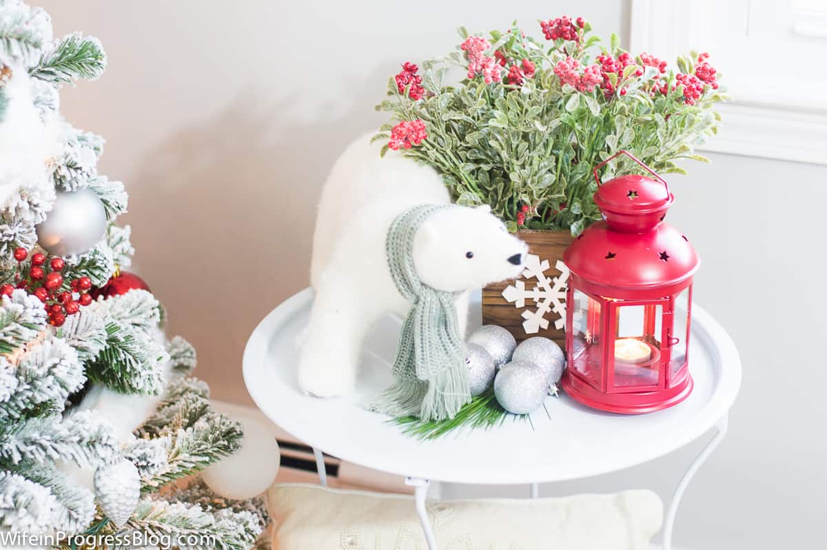 A white side table with a red lantern, a potted plant with red flowers, a polar bear plush and silver ornaments