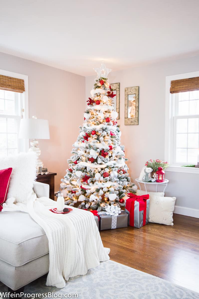 A Christmas tree in the corner of a living room, with red and white decorations, grey gift boxes with red and white ribbon and red and white pillows on a sofa nearby