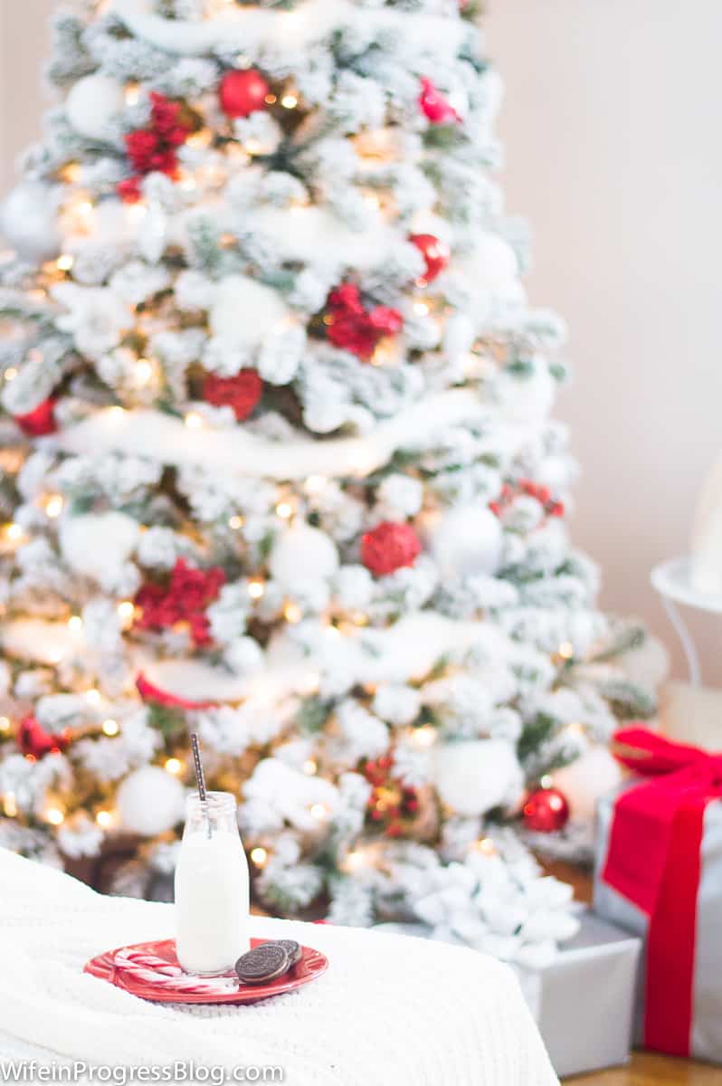 A glass jug of milk, cookies and a candy cane resting on a red plate on a sofa, with the Christmas tree in the background