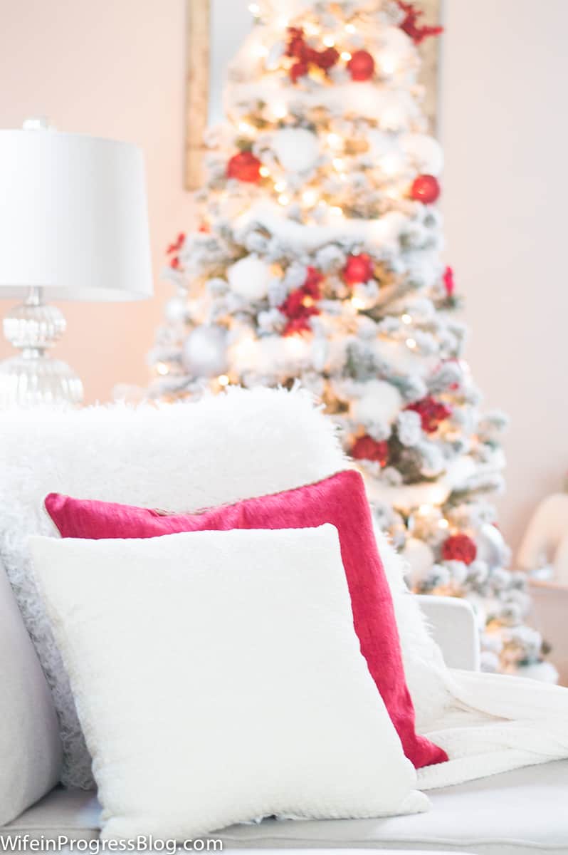 A pair of throw pillows, solid red and white, on a couch in front of a Christmas tree, decorated in red, white and silver
