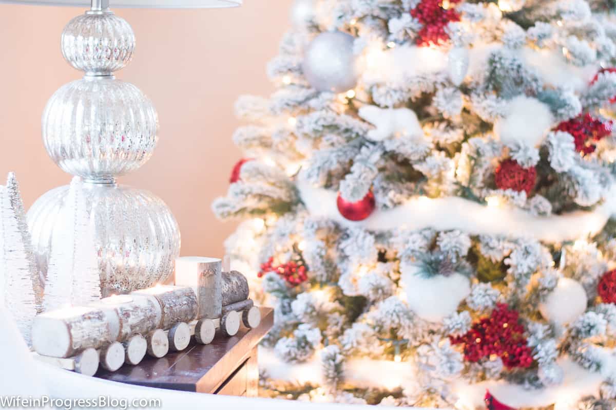 An end table next to a Christmas tree, displaying a silver, wooden train set and a lamp with a decorative glass base
