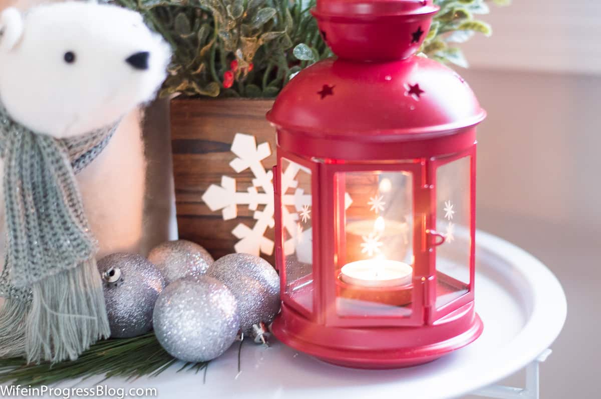 A close-up of a red lantern; silver, glittery ornaments and a potted plant on a white end table