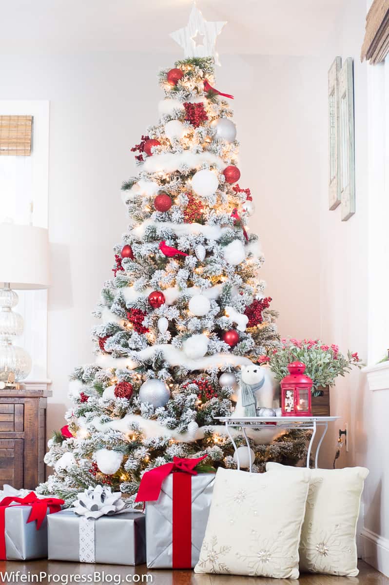 A Christmas tree in the corner, near a side table with a large lamp and another table holding red and white holiday decor