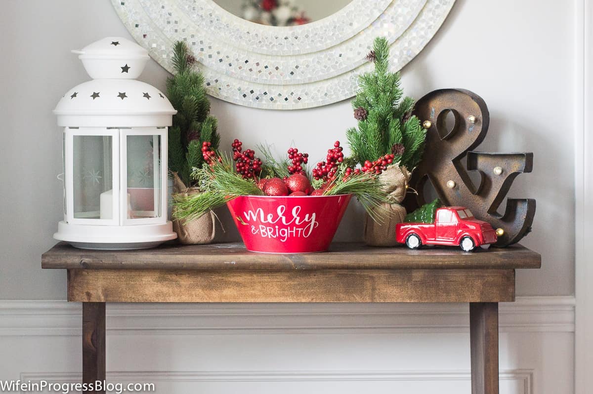 A red bowl with the words \'merry and bright\', on top of a table with other red and white decor, under a round, silver mirror