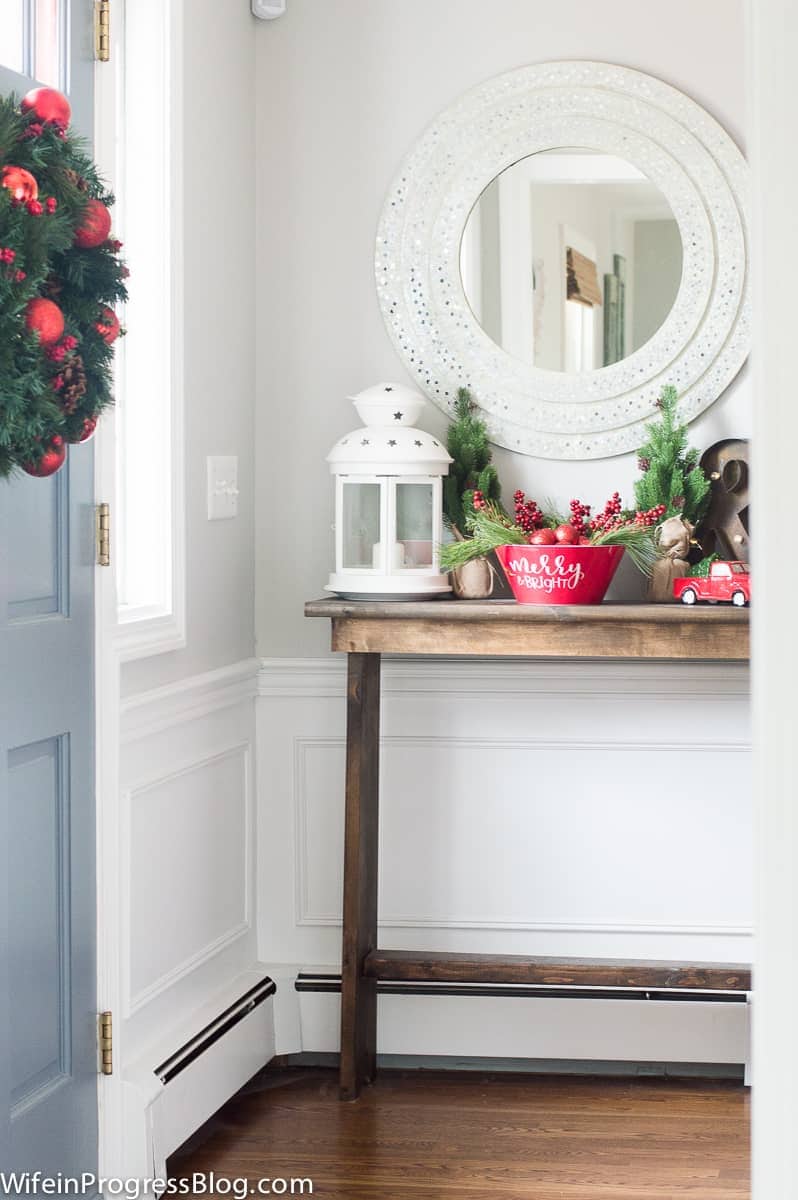A wooden console table holding various red and white holiday decor, near a light blue front door with a large red and green wreath