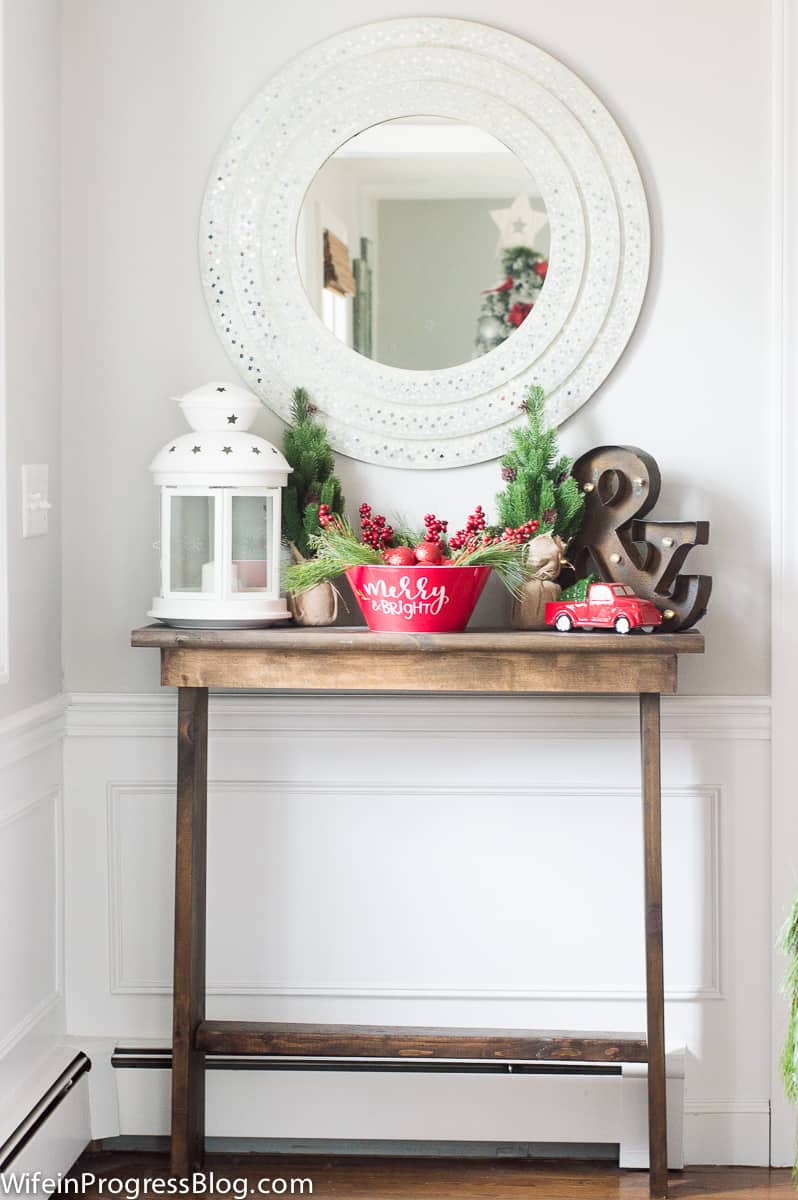 A console table in the entryway, holding a large, white lantern, a red bowl with decorative berries and greenery and various holiday decor