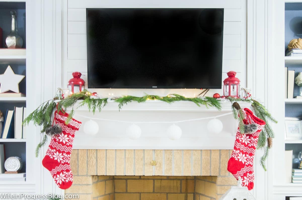 A fireplace mantel with a mantel below the wall-mounted television, holding pine branches, red lanterns and stockings