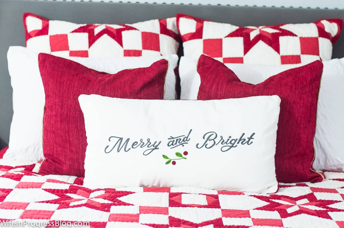 A decorative pillow with the words \'merry and bright\', among solid white and red throw pillows on a red/white patterned bed spread