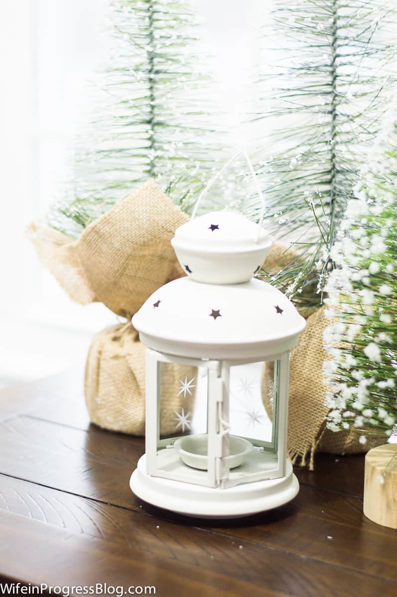 A white lantern with star detailing, in front of artificial pine trees wrapped in burlap