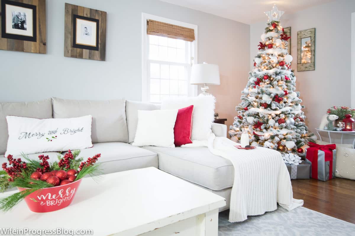 A light grey sofa, with white and red pillows and blanket, near a white coffee table with festive arrangement on top, and Christmas tree in corner